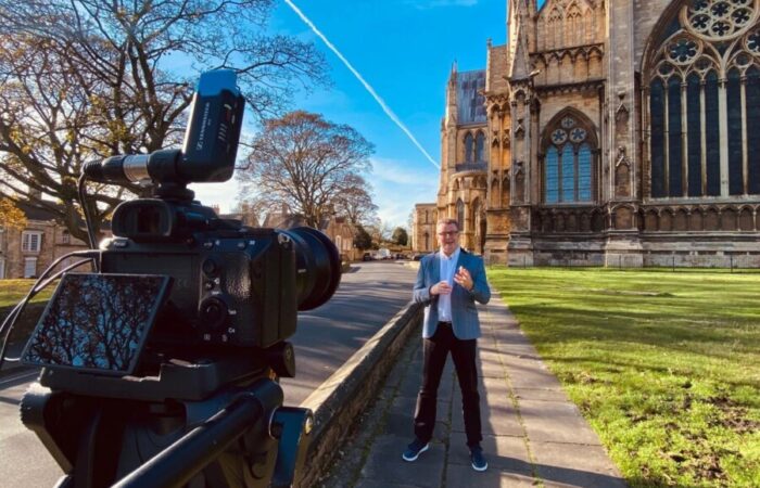 tall man stood outside of Lincoln cathedral in front of a camera